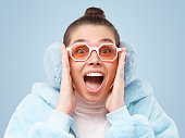 Emotional trendy woman dressed in pink fur coat, wearing earmuffs and glasses, shouting wow