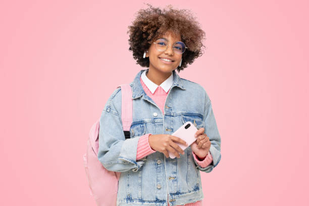 African school girl with afro hairstyle, pink backpack and glasses holding phone African american school girl with afro hairstyle, pink backpack and trendy round glasses holding phone, isolated on pink background cute teen pic stock pictures, royalty-free photos & images