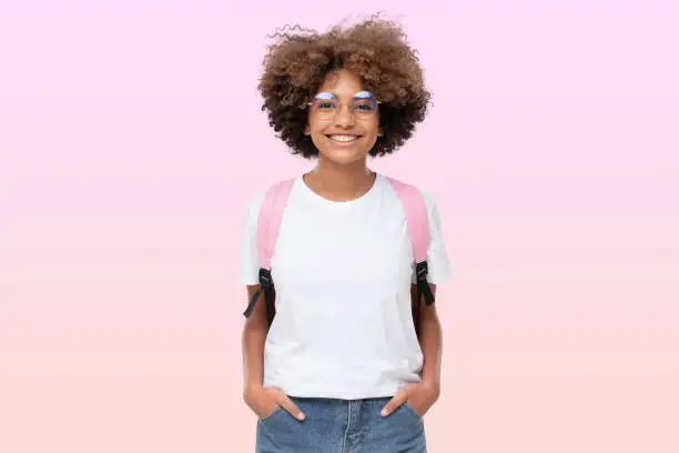 Photo of Portrait of smiling african school girl in white t-shirt, glasses and backpack, isolated on pink