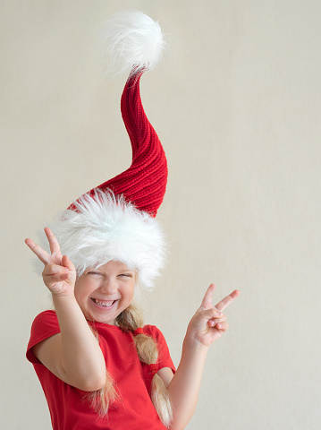 Funny little girl in the Santa hat, standing , smiling and show two Victory with her fingers on the light-brown background