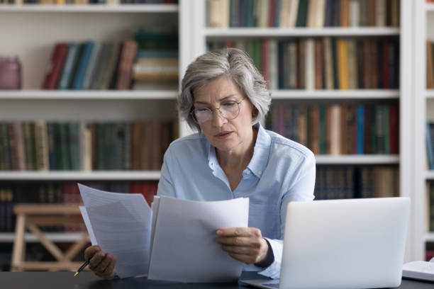 Serious thoughtful middle aged woman worried reading news in document Serious thoughtful middle-aged woman in glasses looks worried read news in formal document sit at workplace desk with wireless computer. Older female review paper letter, learns report feels concerned woman examining stock pictures, royalty-free photos & images