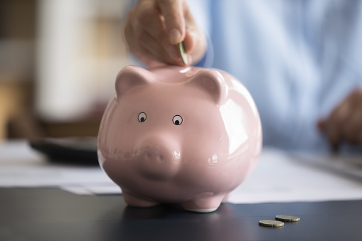 Close up cropped image female hand putting coin in pink piggybank. Save money for future, healthcare medical insurance cover, planning investment, managing household family budget, accounting concept