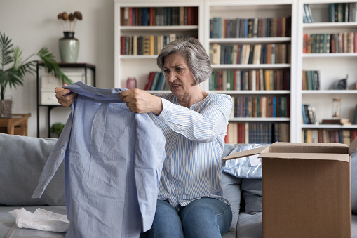 Dissatisfied client of retail services, complains, goods return concept. Aged 60s woman sit on sofa unpack received parcel box check female blouse looks disillusioned by fabric quality, obsolete model