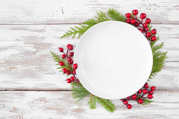 istock Christmas table setting mock up white plate with frame of evergreen branches and red berries on a white wood background 1435623734