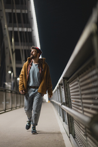 Fashionable young man with a beard walking on a bridge at night. Handsome young man on his way down the bridge alone.
