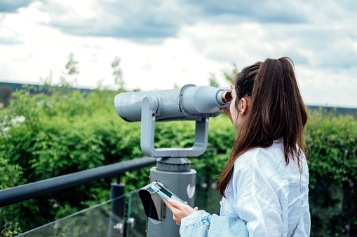 Travel, sightseeing. Young woman tourist looking in binoculars, telescope in the city view. Back view of woman use binoscope in the viewpoint on the river bank.