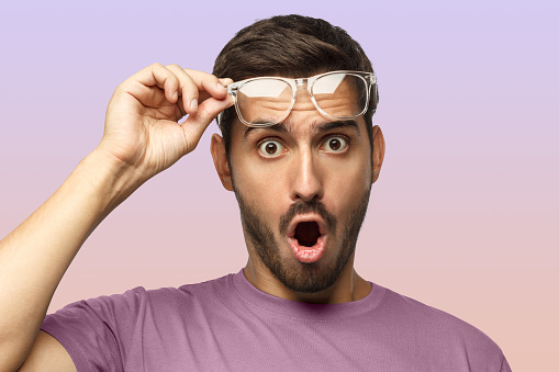 Young man in purple t-shirt shouting OMG with open mouth, surprised by low price and sales, holding transparent glasses