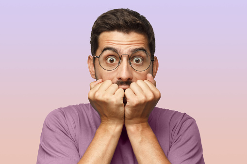 Young man isolated on purple background, covering mouth with hands and round eyes, wearing round eyeglasses, experiencing deep astonishment and fear