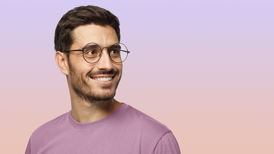 Closeup headshot of young man in round eyeglasses isolated on purple background, smiling happily, looking right, feeling positive, relaxed and joyful