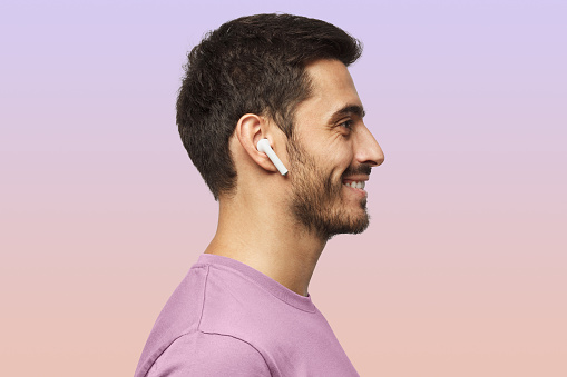 Sideways portrait of smiling young man listening to music or radio, uses modern wireless earphones, wearing purple t-shirt. Copy space for text
