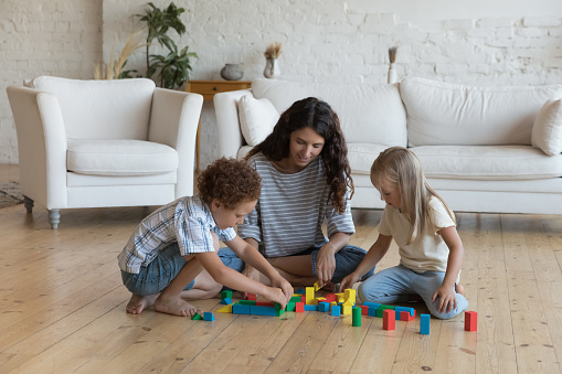 Young mother her children, preschoolers son and daughter sit on warm floor in living room play together colored wooden blocks, spend leisure, carefree weekend at home. Kids development, games concept