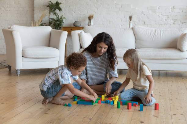 Young mother and children play together colored wooden blocks Young mother her children, preschoolers son and daughter sit on warm floor in living room play together colored wooden blocks, spend leisure, carefree weekend at home. Kids development, games concept nanny stock pictures, royalty-free photos & images