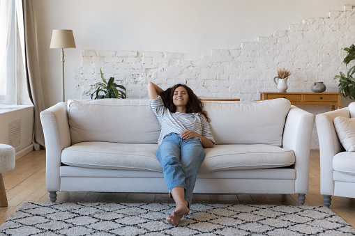 Latina woman daydream relax on sofa at home, housewife accomplish household chores reduces exhaustion, rest alone in living room, full-length. Air condition system inside, stress-free weekend concept