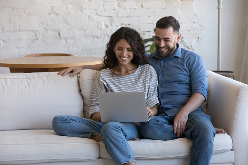 Beautiful millennial couple do e-hopping at home, sit on sofa with laptop smile enjoy distance services for comfort life, make remote order, buy goods feel satisfied. Leisure using modern tech concept