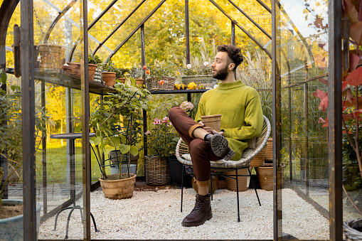 Man sits alone with a flowerpot in greenhouse with plants. Hobby, gardening and solitude concept