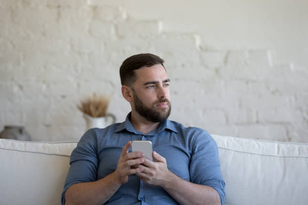 istock Pensive millennial man holds smartphone seated on sofa 1435631340
