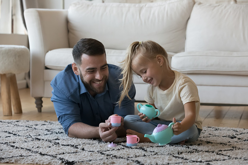 Cute little girl takes care of her loving daddy pours imaginary tea into cup from toy teapot, play together in modern warm living room on weekend use plastic toy dishware. Family playtime, fun concept
