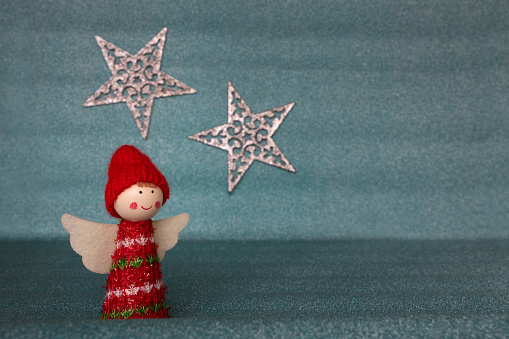 A cute Christmas angel figurine with white wings with a red hat and warm clothes isolated against a blurred glittering pastel blue background with two shining silver stars. Christmas background with empty space for text