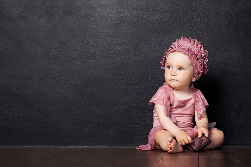 Little girl siting 1 year on a brown wooden floor.  The girl looks aside, holding phone in hand. Thoughtful look. Copy space