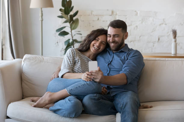 Attractive cheerful couple sit on sofa with cellphone