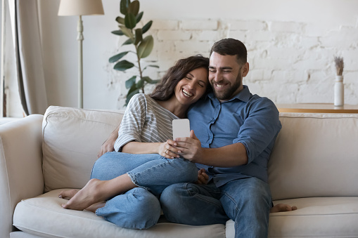 Happy couple sit on sofa with cellphone, make videocall to family, discuss purchase, enjoy new mobile app, consider on-line order. Internet modern tech usage, e-commerce retail services users concept
