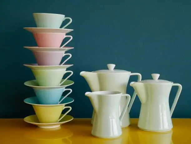 Photo of Stack of colorful vintage tea cups and tea pots on yellow table against petrol background. Afternoon tea party.