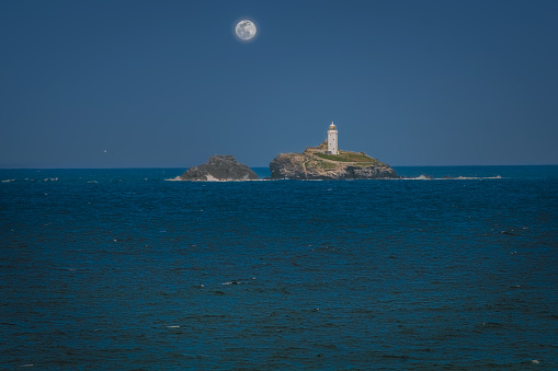 View of lighthouse on rocky outcrop in Cornwall, England,  on clear summer evening; water in foreground and full moon in background
