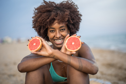 Portrait of a beautiful black woman sitting on a sandy beach in a bikini, smiling and happy, eating grapefruit and enjoying a summer day