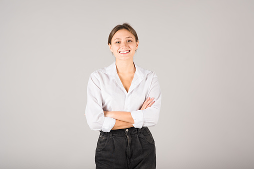 Portrait of a young Caucasian brunette businesswoman in a white shirt with folded arms isolated on a gray studio background.