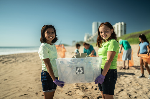 A multi-ethnic group of volunteers picking up garbage, cleaning up the beach. The focus is on a 13 year old African-American girl and her 12 year old friend.