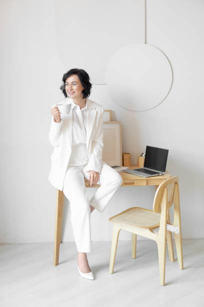 istock Coffee break. A woman in a white suit with a beautiful smile drinks coffee while sitting on a wooden table. Wooden furniture. Bright modern workplace. 1435668901