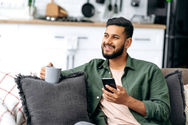 istock Positive indian or arabian guy using his smartphone while sitting on sofa in stylish clothes, browsing internet and social networks, texting with friends or family, found out good news, smiles happily 1435686864