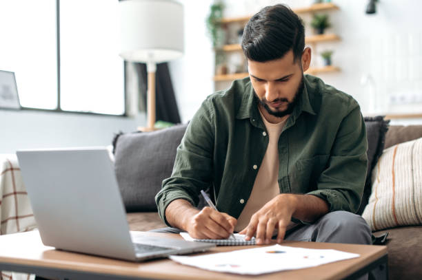 istock Focused smart indian or arabian guy, student or freelancer, sitting at home on sofa in living room, using laptop, working or studying remotely, listening to webinar, taking notes, homework 1435686873