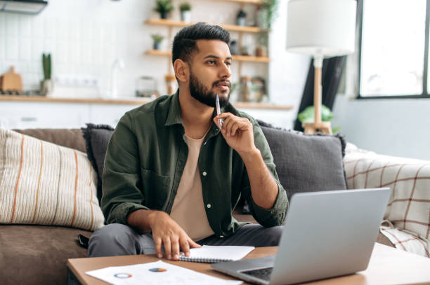 istock Pensive clever indian or arabian guy, student or freelancer, using laptop, working or studying remotely, listening to webinar, taking notes, homework, sits at home on sofa, looks away, thinking 1435686874