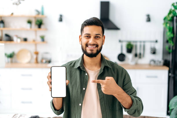 istock Phone screen mockup. Smiling indian or arabian young man, showing smart phone with empty white mock-up screen, copy-space for advertising or presentation, standing in living room, looks at camera 1435686912