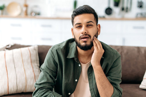 Frustrated unhealthy indian or arabian man in casual clothes, pressing cheek with hand, suffering toothache, caries, sits on a sofa in living room, needs the help of a dentist. Dental problems concept