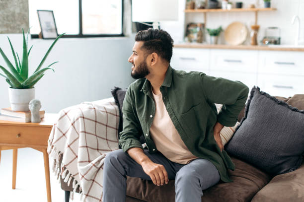 istock Problems with the back, spine, sciatica. Sad Arabian or Indian man, dressed in casual wear, sitting on sofa in living room, experiencing acute pain in the back, lower back, has chronic joint problems 1435686929