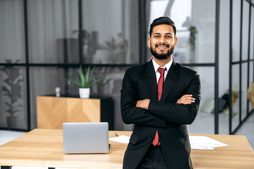 Positive handsome confident successful Indian or Arabian businessman, in a suit, male entrepreneur, stands in a modern office near the desktop with crossed arms, looks at the camera, smiles friendly