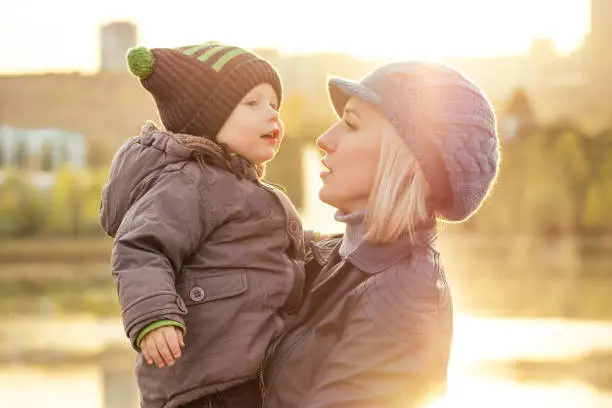 Mother in a warm jacket and knitted hat hugging her baby boy walking in the autumn park. stylish blondewoman with a child against the backdrop of the lake