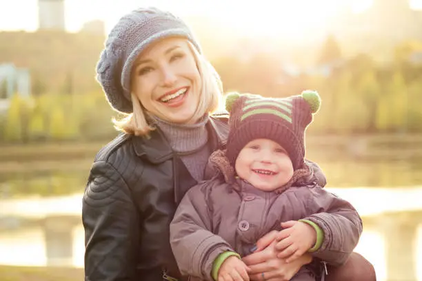 Mother in a warm jacket and knitted hat hugging her baby boy walking in the autumn park. stylish blondewoman with a child against the backdrop of the lake.
