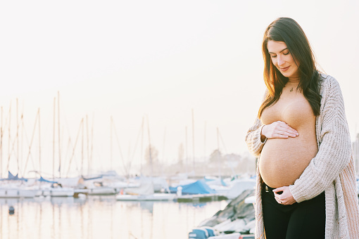 Outdoor portrait of beautiful pregnant woman touching belly, early morning walk next to lake