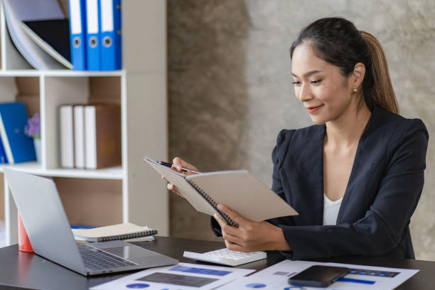 istock Asian businesswoman sitting at work using a calculator to calculate analytical statistics front view of beautiful woman holding pen and graph paper with laptop on desk in office 1435723362