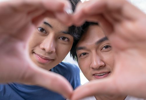Young Asian gay couple in a romantic mood making heart-shaped hand gesture. LGBT male couple showing love and affection while playing with camera in home setting.