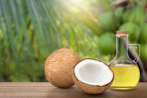 Coconut oil in glass bottle and brown coconut fruit on wooden table with coconut tree background.