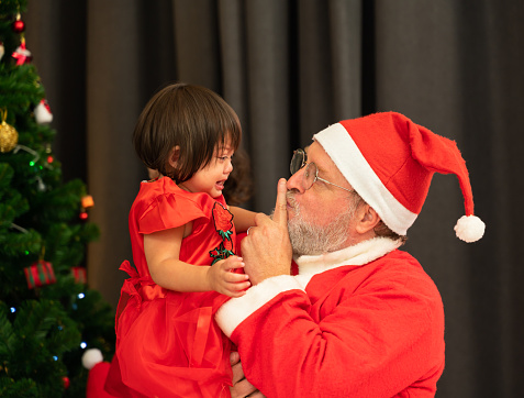 Santa Claus wearing red costume keeping forefinger by his mouth while trying to comfort shouting crying toddler girl near Christmas tree in living room at home