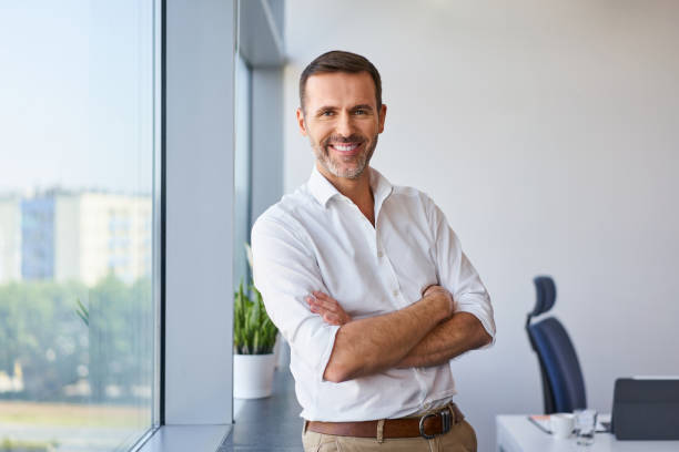 портрет улыбающегося среднего взрослого бизнесмена, стоящего в корпоративном офисе - бизнесмен стоковые фото и изображения