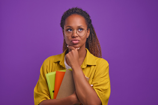 Young smart pensive African American business woman dressed in casual style with notepads or diaries in hand touching chin and pouting lips coming up with new idea, stands posing in purple studio