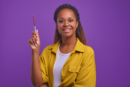 Young independent successful African American woman with big smile raises hand with pen wants to speak or report new idea for starting startup or new company stands on purple background. Casual girl