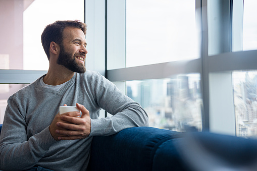 Mid-shot portrait of handsome Latin American man young man looking out the window with cup of tea in hand