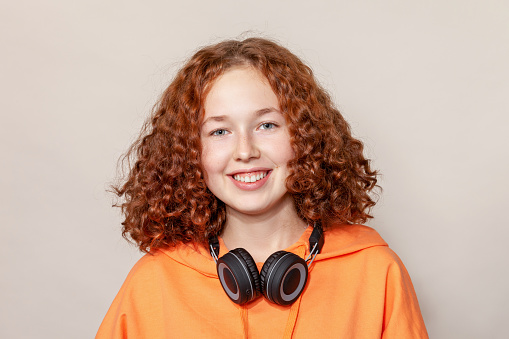 Close-up studio portrait of a cheerful red-haired teenage girl in an orange hooded shirt with headphones on a beige background